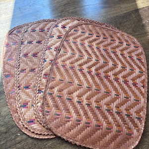 May include: A set of four woven placemats. Each placemat has a rectangular shape with rounded corners and a decorative border. The mats are made of natural-colored woven material with a colorful geometric pattern.