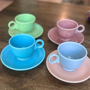 Op de afbeelding: Een set van vier keramische theekopjes met bijpassende schoteltjes in pasteltinten, waaronder lichtgroen, oudroze, fel turquoise en zacht korenbloemblauw. Elk kopje heeft een rond oortje en horizontale ribbels aan de buitenzijde.