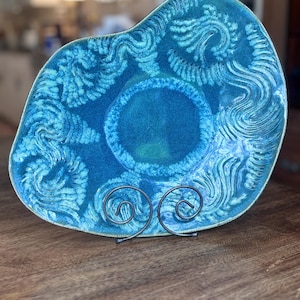 Peut inclure: Une assiette décorative en céramique de forme libre dans les tons de bleu, posée sur un support en métal. L'assiette présente une surface texturée avec des motifs tourbillonnants et un cercle central bleu plus foncé. L'assiette est exposée sur une surface en bois.