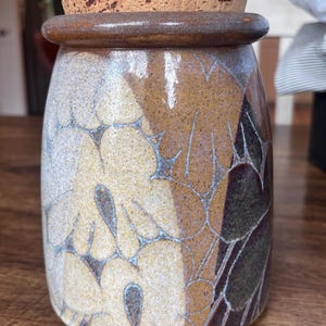 May include: A cylindrical ceramic storage jar with a speckled finish in shades of cream, tan, and dark brown. The jar features a repeating floral pattern outlined in thin lines and is topped with a round, natural-colored cork stopper lid.