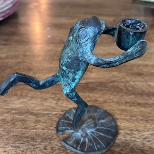 May include: A bronze-toned metal candle holder shaped like a frog standing on a lily pad base. The frog is depicted in a leaping pose, holding a small cylindrical cup for a candle in its front limbs. The metal has a weathered, dark green patina.
