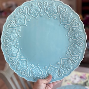 May include: A round, light blue ceramic plate featuring a scalloped edge and an intricate, raised relief pattern of stylised floral and leaf motifs encircling the smooth centre. The glaze has a subtle crackle finish throughout the surface.