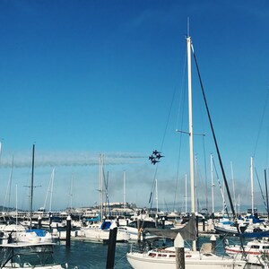 May include: A sunny day at a marina filled with sailboats, with a squadron of blue and white jets flying overhead. The clear blue sky and calm water create a picturesque scene, with a distant view of a city.