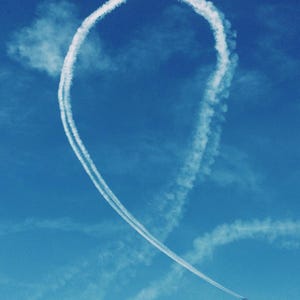 May include: A bright blue sky with a white contrail forming a large loop, resembling a question mark or a stylized heart. A distant airplane is visible at the bottom, leaving a white trail. The image captures an aerial display.