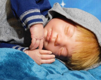 May include: A sleeping baby doll wearing a gray hooded sweatshirt with blue sleeves and a blue blanket. The doll has blonde hair and is lying on its side.