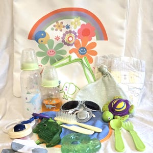 Puede incluir: Una colección de artículos para bebés, que incluye biberones, un babero, un chupete, gafas de sol y una tortuga de juguete. Una bolsa tote blanca con un diseño de arcoíris y flores está en el fondo. Otros artículos incluyen un pañal, calcetines y un gorro de bebé.