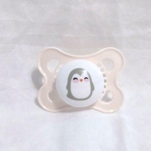 Peut inclure: Une sucette blanche avec un motif de pingouin gris. Le pingouin a un bec rose et des joues roses.