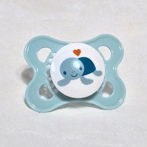 Könnte beinhalten: Ein hellblauer Babyschnuller mit einem runden, weißen Mittelknopf. Der Knopf zeigt eine grafische Illustration einer lächelnden hellblauen Schildkröte mit einem dunkelblauen Panzer und einem kleinen orangefarbenen Herz über dem Kopf.