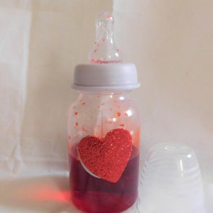May include: A clear baby bottle with a red glitter heart sticker on the side. The bottle is filled with a red liquid. The bottle has a white cap and a clear nipple.