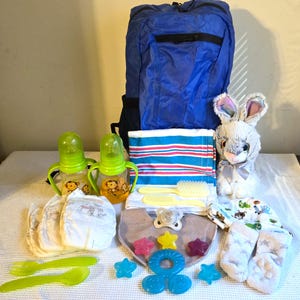 May include: A blue backpack containing baby supplies including two green sippy cups with monkey graphics, a striped blanket, a plush rabbit, diapers, a bib, a pacifier, star-shaped teethers, a blue ring teether, a hairbrush, a comb, and socks.