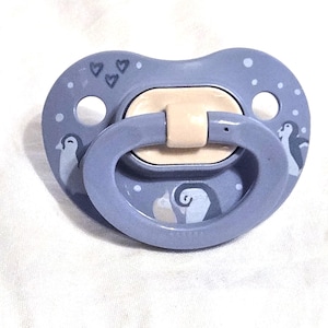 May include: A baby's soother in a soft blue colour, decorated with penguin illustrations and small heart shapes. The soother has a light beige teat and a matching blue handle, designed for infants.