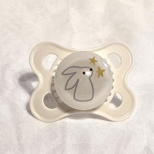 May include: A cream-colored baby pacifier with a bunny and star design. The pacifier has a rounded nipple and a shield with two holes. The center of the pacifier features a gray bunny and two gold stars.