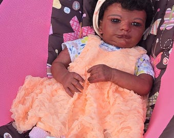 May include: A realistic baby doll with dark skin and curly black hair, wearing a peach-coloured ruffled dress, a floral patterned shirt, a cream-coloured headband with a bow, light pink socks, and white shoes with pink ribbon accents.