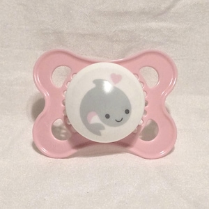 Peut inclure: Une sucette rose pour bébé avec un centre rond et blanc représentant une illustration de baleine grise et un petit cœur rose. La sucette a un bouclier en forme de papillon avec deux trous de chaque côté.