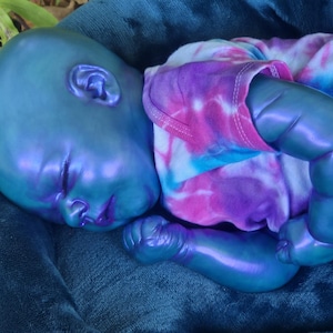 Puede incluir: Una muñeca bebé de tono azulado con un acabado brillante, acostada sobre una superficie azul oscuro. La muñeca lleva una camiseta teñida con tintes de rosa, morado y azul. La piel del bebé tiene un brillo iridiscente sutil.