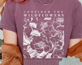 May include: Berry-colored t-shirt with the text "CONSIDER THE WILDFLOWERS" above a floral illustration. The shirt also features a Bible verse. A "MORE COLORS" sticker is visible.