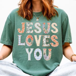 Puede incluir: Camiseta verde claro con el texto "JESUS LOVES YOU" en un diseño de patchwork. Las letras están en varias telas florales y estampadas. La modelo lleva vaqueros de lavado claro y está sentada con las piernas cruzadas.