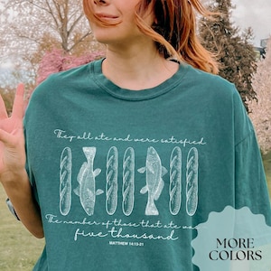 May include: Teal green t-shirt with a white graphic of fish and loaves of bread. The text on the shirt reads "They all ate and were satisfied. The number of those that ate was five thousand. Matthew 14:19-21."