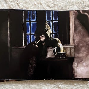 May include: A print depicting a figure in a hooded cloak smoking a pipe, seated at a table. A window with blue light is behind the person. A candle and a mug are on the table. The image has a dark, moody atmosphere.