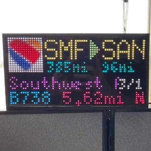 Puede incluir: Una pantalla LED negra muestra información de vuelos. El logotipo de Southwest Airlines está en la esquina superior izquierda. El texto incluye "SMF a SAN", detalles del vuelo y la distancia en millas. La pantalla está iluminada con varios colores.