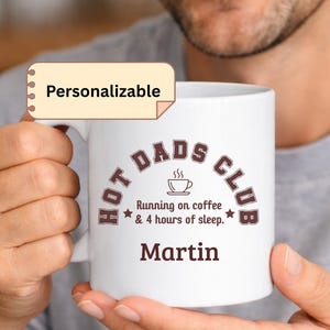 Könnte beinhalten: Weiße Keramik-Tasse mit dem Text "HOT DADS CLUB" in Braun, zusammen mit "Running on coffee & 4 hours of sleep." und dem Namen "Martin". Die Tasse wird von einer Person gehalten.