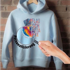 May include: Light blue hoodie with a colorful dog graphic and the text "FLAUSCH ATTA CKE". A magnifying glass shows the word "personalisierung" and "Dogs name". The hoodie is displayed on a wooden hanger against a brick wall.