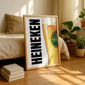 Puede incluir: Un p&oacute;ster enmarcado con un primer plano de un vaso de cerveza y el logotipo de Heineken. La palabra "HEINEKEN" est&aacute; impresa verticalmente en letras negras en negrita. El p&oacute;ster est&aacute; en un marco de madera clara.