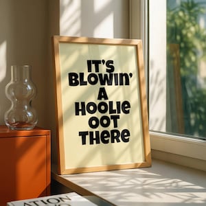 Peut inclure: Affiche encadrée avec le texte "IT'S BLOWIN' A HOOLIE OOT THERE" en noir, en gras. L'affiche est dans un cadre en bois clair et est appuyée contre une fenêtre. Un vase en verre transparent et un meuble orange sont également visibles.