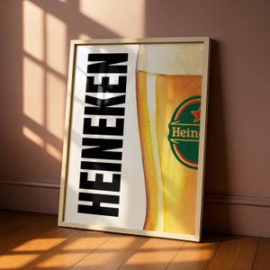 Puede incluir: Impresi&oacute;n enmarcada con un vaso de cerveza y el logotipo de Heineken. La palabra "HEINEKEN" est&aacute; impresa verticalmente en letras negras en negrita. El vaso de cerveza est&aacute; lleno de un l&iacute;quido dorado. El marco es de madera.