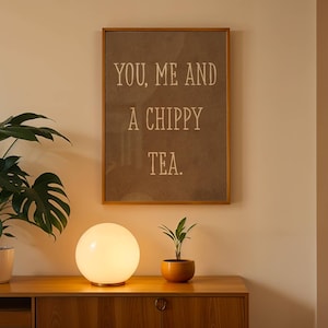 May include: A rectangular wall art piece in a wooden frame featuring the text YOU, ME AND A CHIPPY TEA. in white lettering on a brown background. The artwork is displayed above a wooden sideboard with a spherical white lamp and two potted plants.