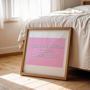 Puede incluir: Una impresión enmarcada con el texto "A dinnae rise and shine, a wake up, and greet aboot it" en un marco de madera clara. La impresión tiene un fondo rosa y está apoyada contra una cama en un dormitorio.