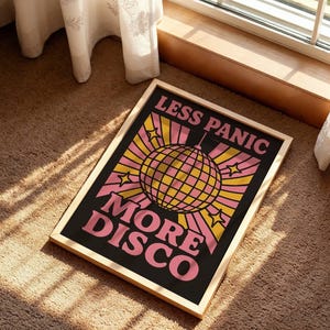 Könnte beinhalten: Gerahmtes Poster mit den Worten "LESS PANIC MORE DISCO" in rosa Schrift. Das Poster zeigt eine Discokugel-Grafik in Gelb und Rosa auf schwarzem Hintergrund. Der Rahmen hat eine helle Holzfarbe.