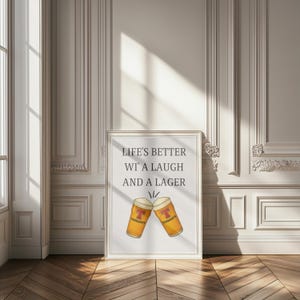 May include: Framed print with the text "Life's better wi' a laugh and a lager" above two cartoon beer glasses. The glasses are orange with the Tennent's logo. The print is in a white frame, set against a white wall.