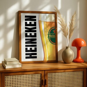 Puede incluir: Impresi&oacute;n enmarcada con el logotipo de Heineken y un vaso de cerveza parcialmente visible. La palabra "HEINEKEN" est&aacute; impresa verticalmente en negro. La impresi&oacute;n se muestra en un mueble de madera con elementos decorativos.