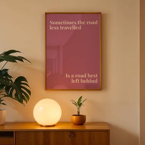 May include: A framed print with the text "Sometimes the road less travelled Is a road best left behind" hangs on a wall. A glowing globe lamp and potted plants sit on a wooden cabinet below the print.