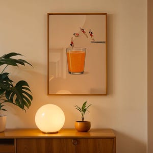 May include: A framed art print shows divers leaping into a large glass of orange juice. The artwork has a light background and a wooden frame. The print is displayed above a wooden cabinet with a round, glowing lamp and a potted plant. The lamp is 25 cm in diameter.