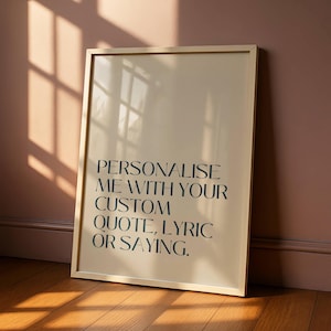 以下が含まれることがあります： 「PERSONALISE ME WITH YOUR CUSTOM QUOTE, LYRIC OR SAYING」のテキストが入ったフレーム付きプリント。プリントは薄いベージュの背景と金のフレームです。画像は木の床のある壁に立てかけられています。