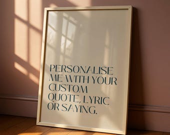 May include: A framed print with the text "PERSONALISE ME WITH YOUR CUSTOM QUOTE, LYRIC OR SAYING." The print has a light beige background and a gold frame. The image is leaning against a wall with a wooden floor.
