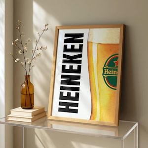 Puede incluir: Un cartel enmarcado con el logotipo de Heineken y una imagen parcial de un vaso de cerveza. La palabra "HEINEKEN" est&aacute; impresa verticalmente en letras negras en negrita. El vaso de cerveza tiene espuma y un logotipo verde y rojo.