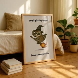 May include: A framed print featuring a cartoon raccoon wearing a witch hat and riding a broom. The raccoon is brown, gray, and white, with a black hat. The text on the print reads "people pleasing is over" and "become unacceptable."
