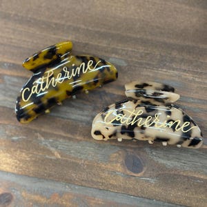 May include: Two hair claw clips with the name "Catherine" in gold script. One clip has a tortoise shell pattern in shades of brown and black. The other clip has a beige and black pattern. Both clips are on a wooden surface.