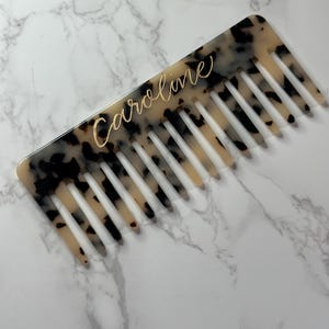 May include: A tortoiseshell-patterned comb with the name "Caroline" in gold script. The comb has wide teeth and a rectangular shape. The background is a white marble surface.