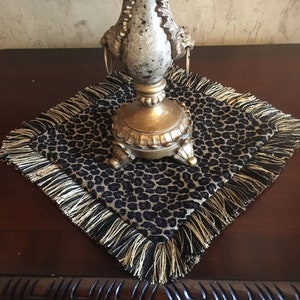 May include: A decorative table square with a black and tan leopard print pattern, trimmed with gold fringe. A gold-colored, ornate lamp base sits atop the square. The square is placed on a dark brown wooden surface.