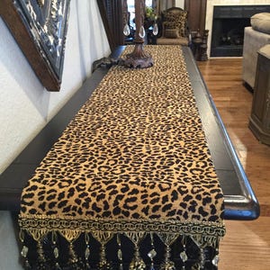 May include: A leopard print table runner with gold and black fringe and beaded accents. The runner is on a dark wood table.