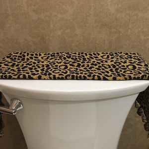 May include: A leopard print toilet tank cover with gold tassels. The cover is made of a soft, plush fabric and is designed to add a touch of style to any bathroom.