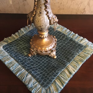 May include: A decorative gold-toned metal candlestick with a detailed base and ornate design, resting on a square teal placemat with a textured pattern and fringed edges. The candlestick has a speckled, aged appearance.