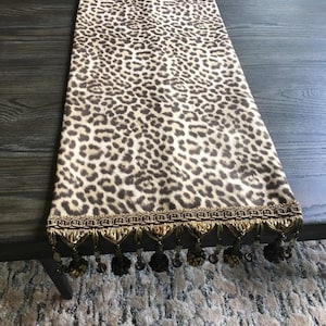 May include: A leopard print table runner with a brown and tan spotted pattern. The runner has a decorative gold trim with hanging tassels. The runner is displayed on a dark wooden table.