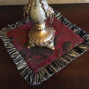 May include: A red and gold patterned square table runner with black and gold fringe. The runner is on a dark wood surface and a gold decorative vase is on top of the runner.
