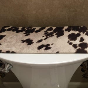 May include: A white toilet bowl with a brown and white cowhide patterned toilet seat cover with tassels.