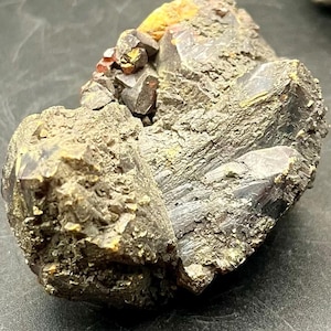 May include: A close-up of a rough, dark-coloured mineral specimen. The stone has a textured surface with a mix of dark grey, brown, and yellow hues. Small, reddish-orange crystals are visible on the top of the mineral.